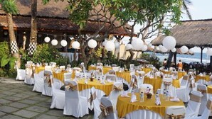 An outdoor wedding reception with round tables covered in white tablecloths and yellow centerpieces, set against a backdrop of a beach and thatched-roof structures.
