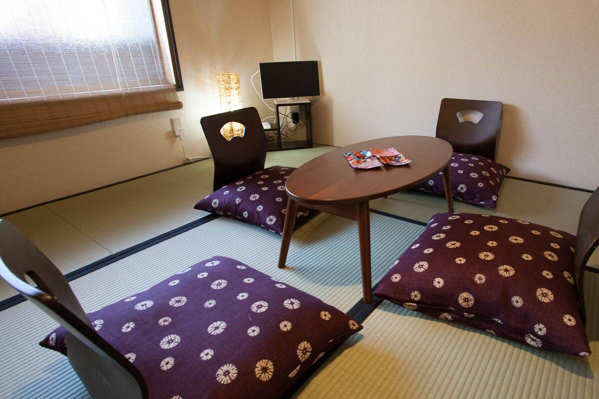 A tatami mat room with a low table and chairs, a television, and a window with blinds.