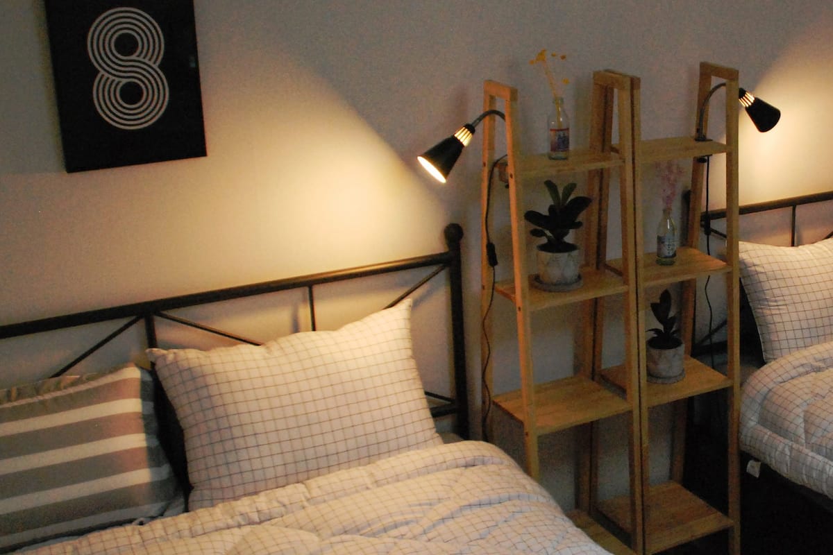 Quarto com duas camas, cada uma com cabeceira, uma estante com plantas e uma luminária na parede.