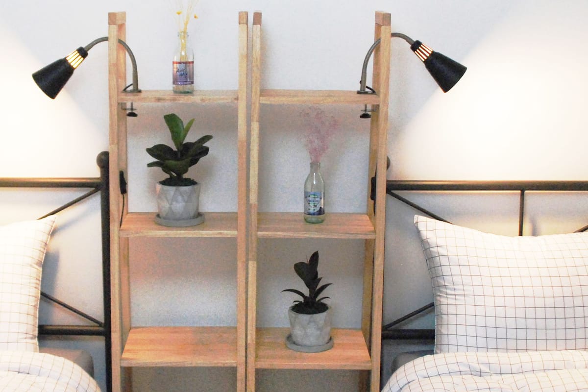 Quarto com duas camas, cada uma com almofadas listradas, uma prateleira de madeira com plantas e uma luminária fixada na parede.