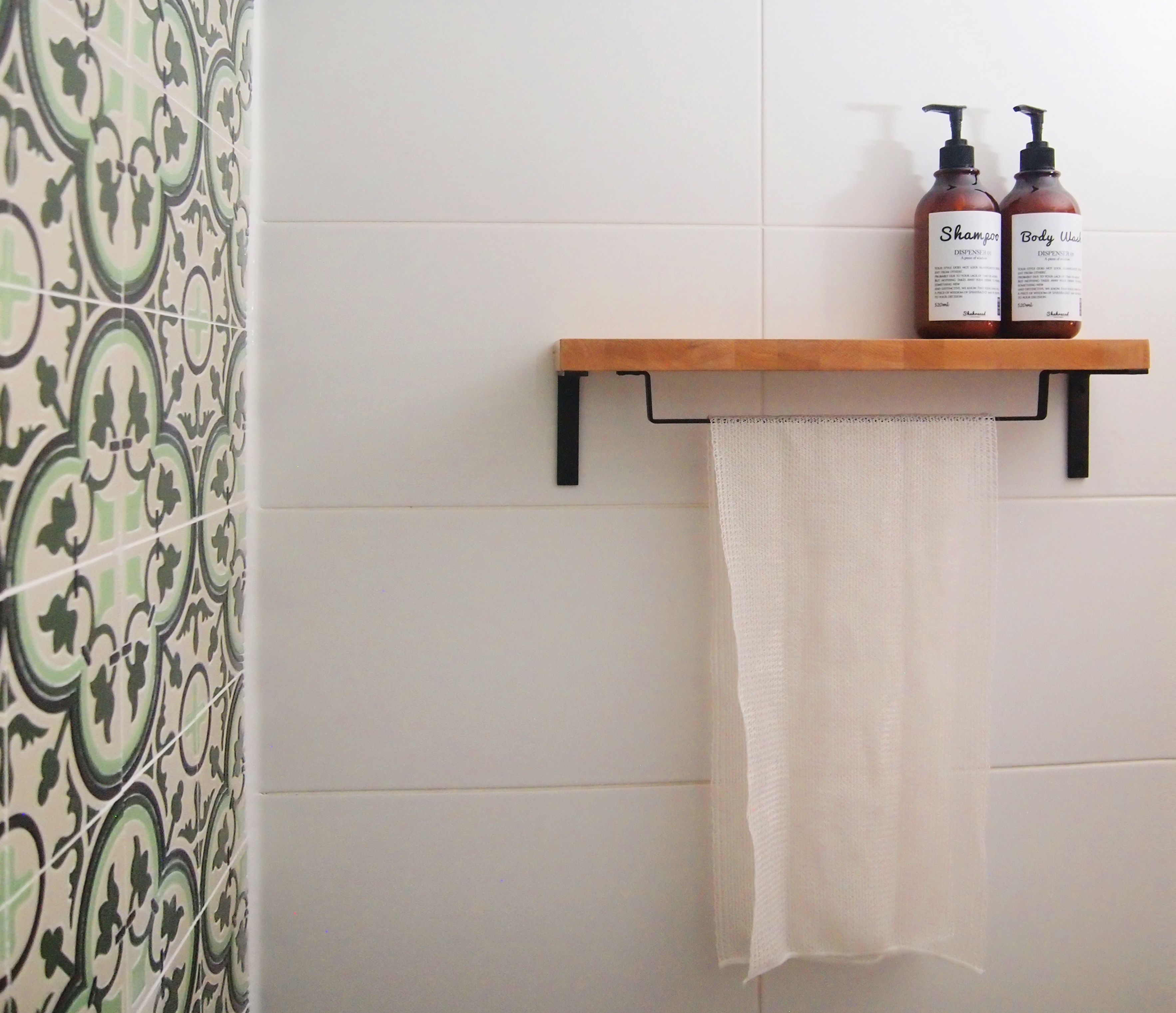 A bathroom with a patterned tile wall, a wooden shelf holding two bottles of shampoo and body wash, and a white towel hanging on a metal rack.