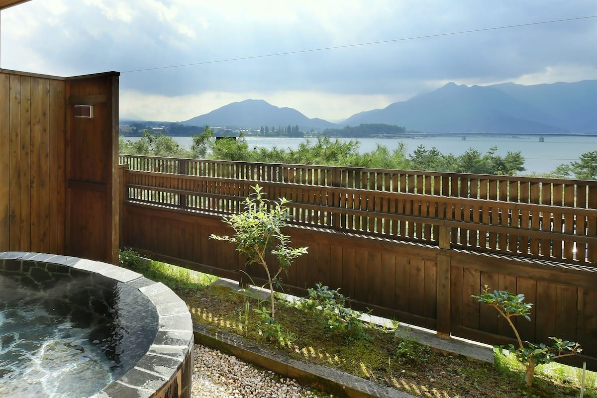 A hot tub with a stone edge, a wooden deck, and a view of a lake and mountains.