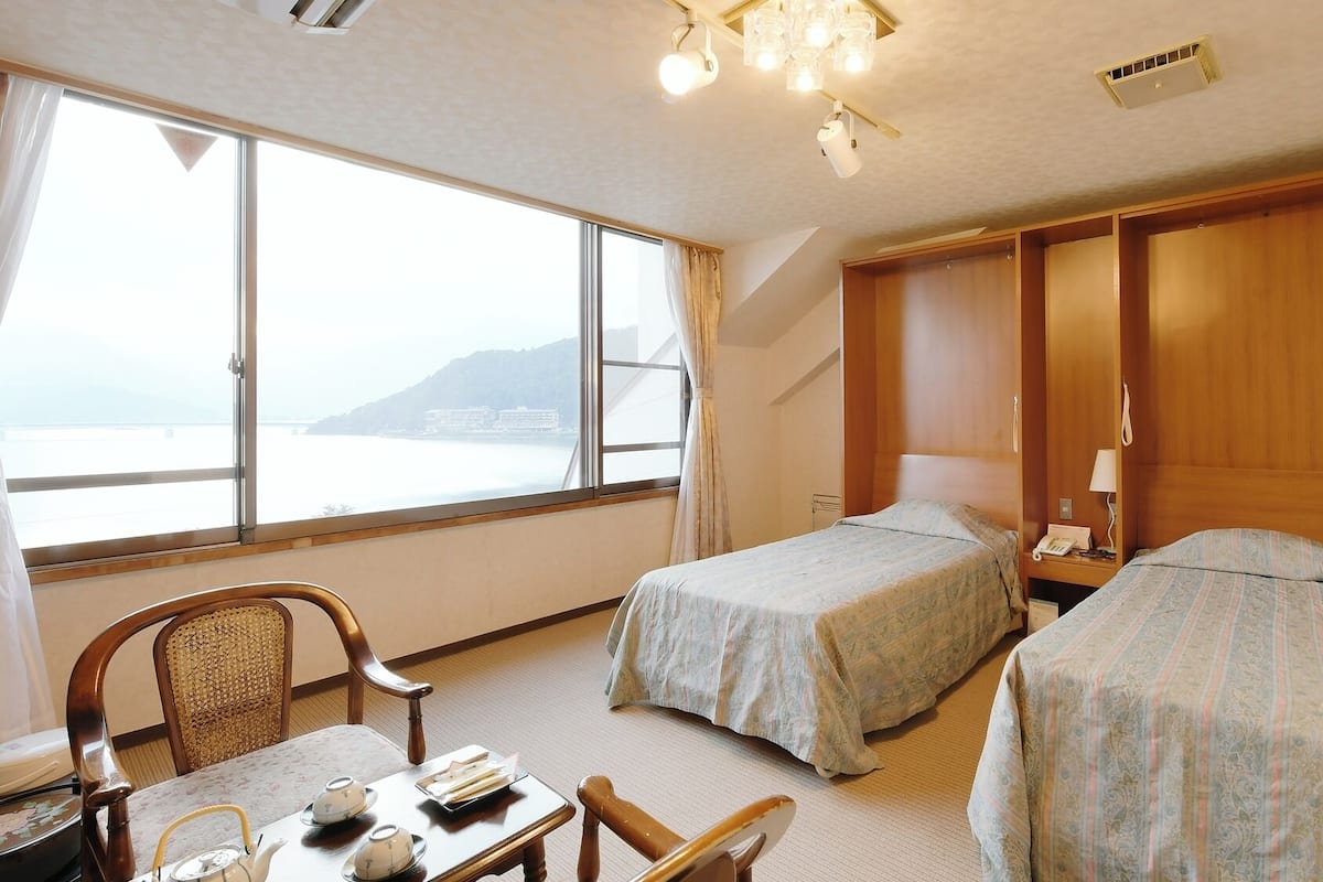 A hotel room with two beds, a desk with a chair, a table with a teapot, and a view of the sea.
