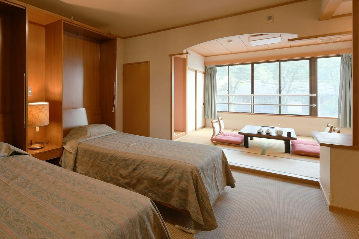 A hotel room with two beds, a bedside table with a lamp, a window with a view, and a small seating area with a table and chairs.