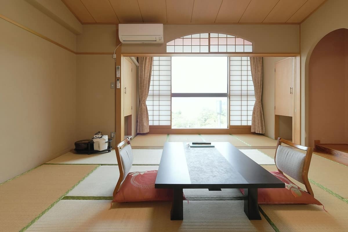 A traditional Japanese room with tatami flooring, a low table, sliding doors, and a view through a window with a grid pattern.