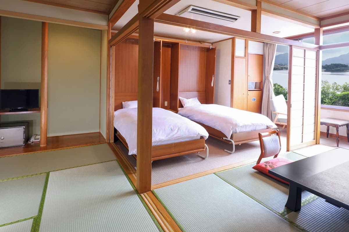 A traditional Japanese-style room with tatami flooring, two single beds, a wooden wardrobe, a television, and a view of a lake.