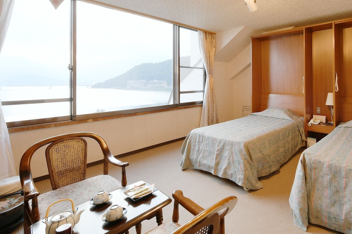A hotel room with two beds, a wooden wardrobe, a table with a teapot and cups, and a view of the sea.