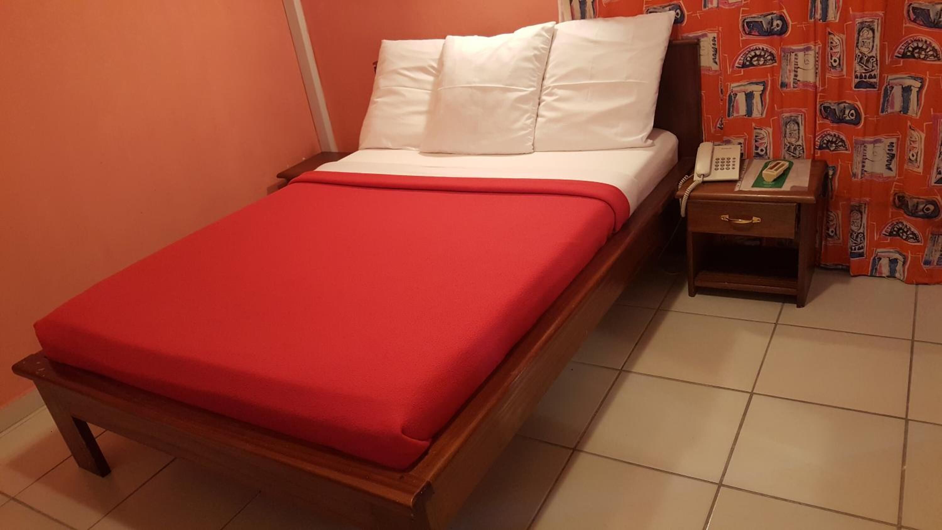 Un llit individual amb un estovat vermell i almoadines blancs en una habitació amb parets taronges i una cortina amb dibuixos.
