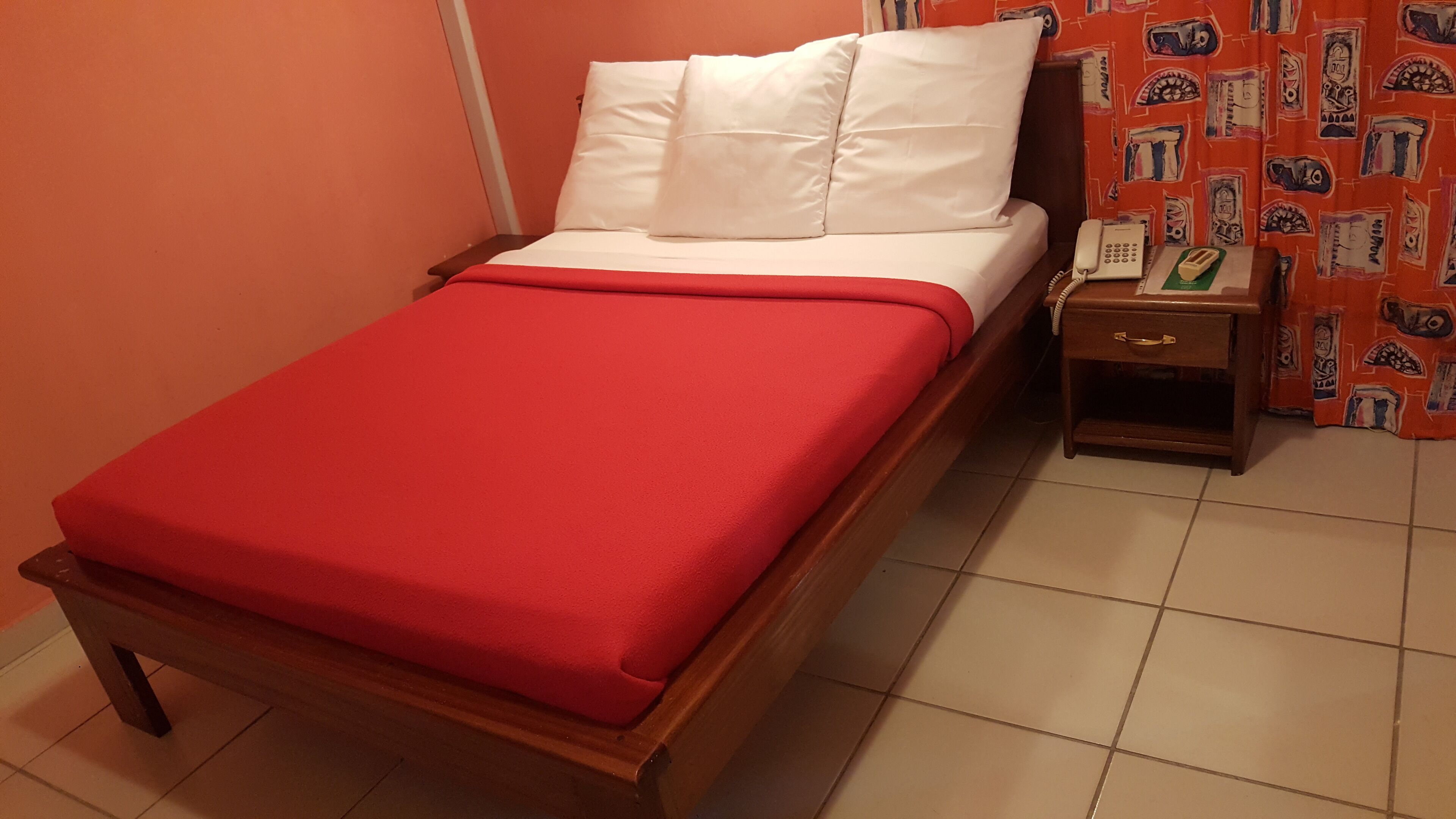 Un lit simple avec une couverture rouge, des oreillers blancs, une tête de lit en bois, une table de chevet avec un téléphone et un grille-pain, et un rideau à motifs.