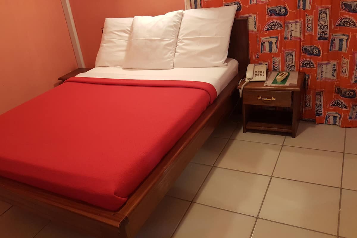 Een enkel bed met een rode deken, witte kussens, een houten hoofdbord, een nachtkastje met een telefoon en een broodrooster, en een gordijn met een patroon.