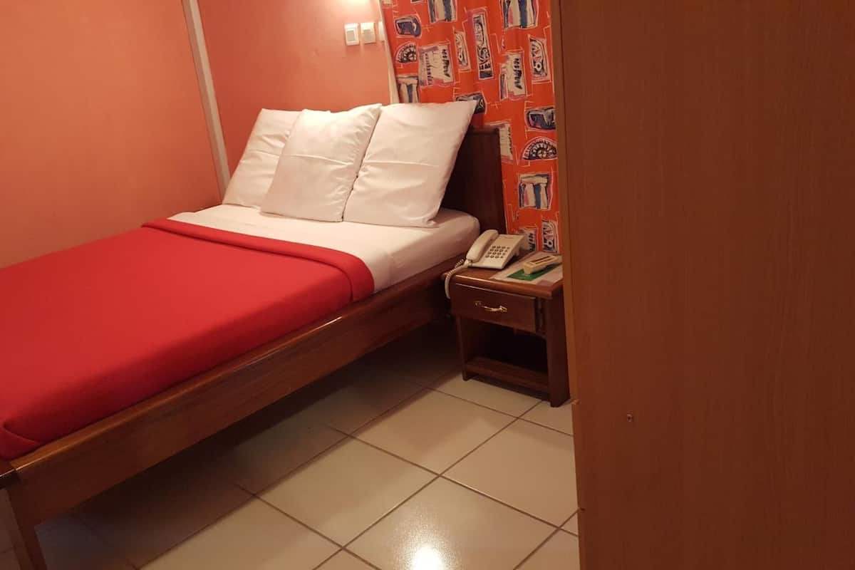Een eenpersoonskamer met een rood sprei, witte kussens, een houten nachtkastje met telefoon, en een behangen wand.
