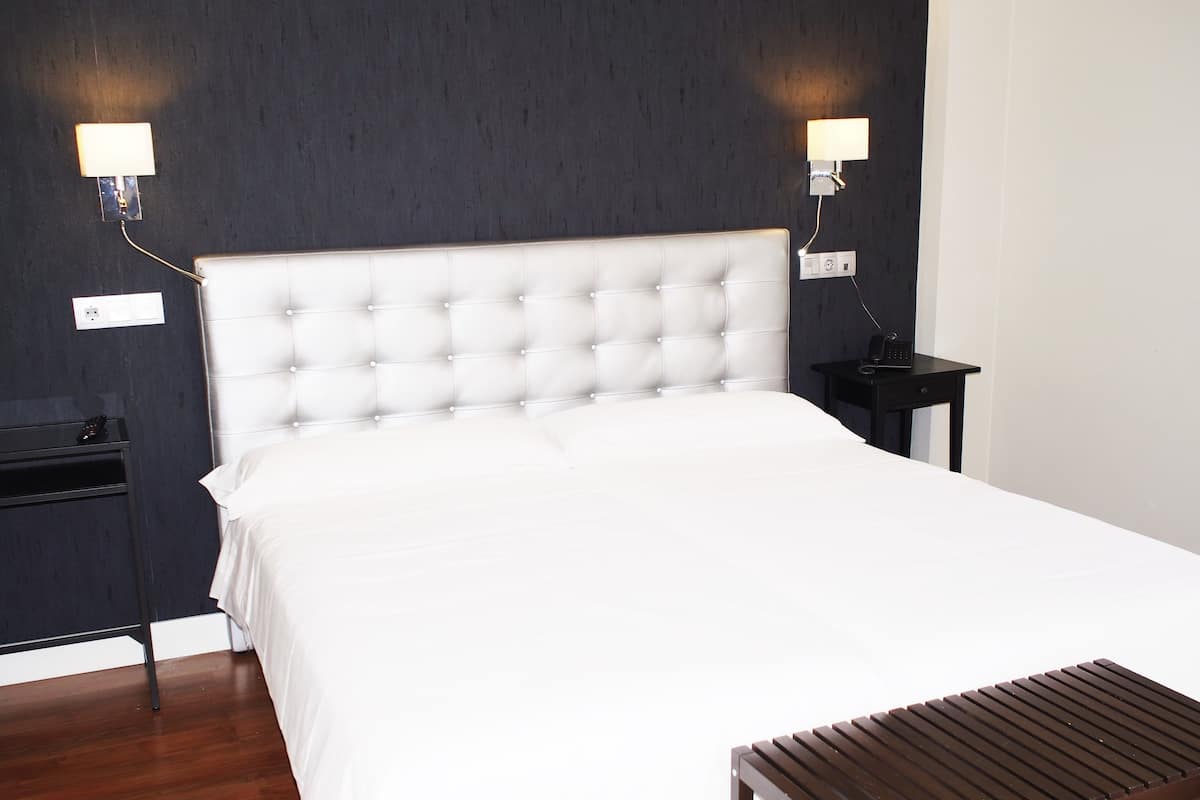 A neatly made bed with a tufted headboard, two bedside tables with lamps, and a wooden bench in a modern room with dark walls.