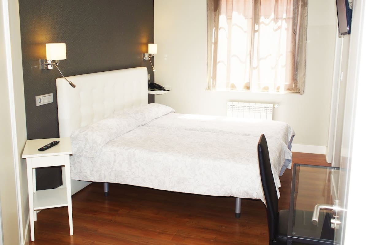 A hotel room with a bed, bedside tables, a desk, a chair, a TV, and a window with curtains.