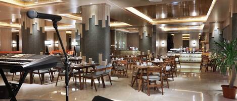 A spacious hotel lobby with a modern design, featuring a grand piano, a stage with a microphone, and a large seating area with tables and chairs.