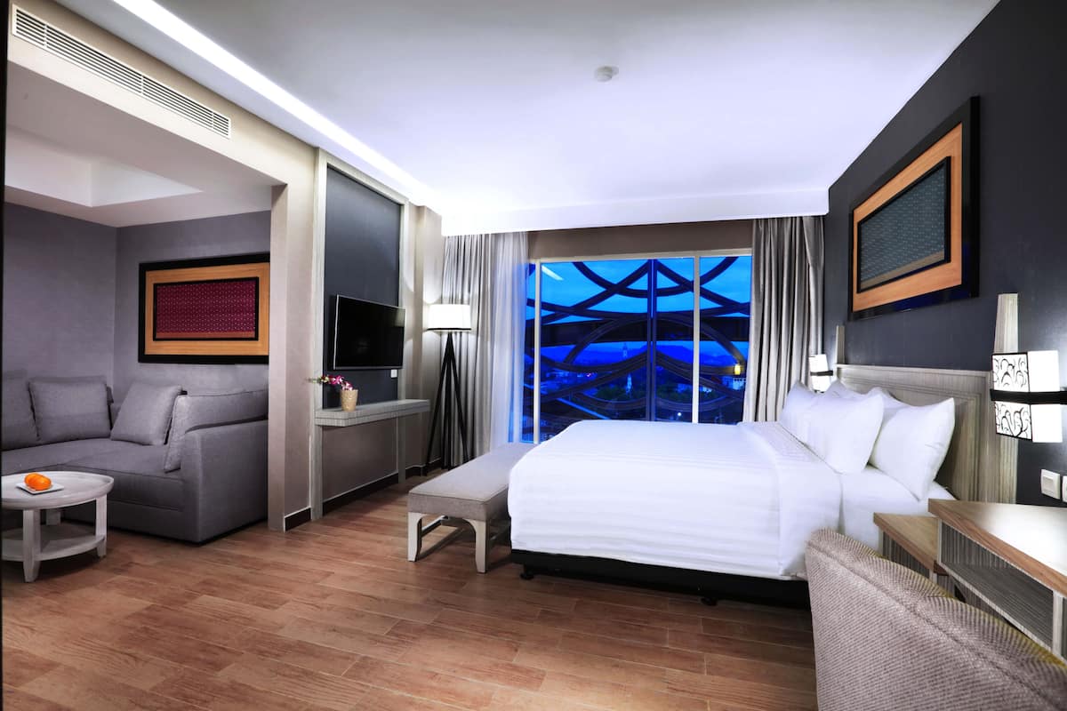 A modern hotel room with a large bed, a sofa, a small table with an orange, and a view of a blue-lit structure outside.