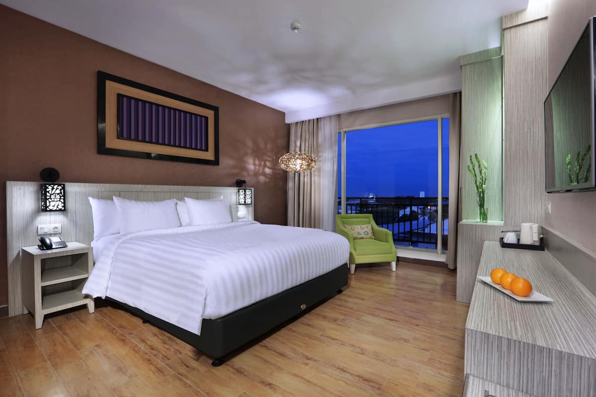 A modern hotel room with a large bed, bedside tables, a green armchair, a small table with oranges, and a view of the city at night.