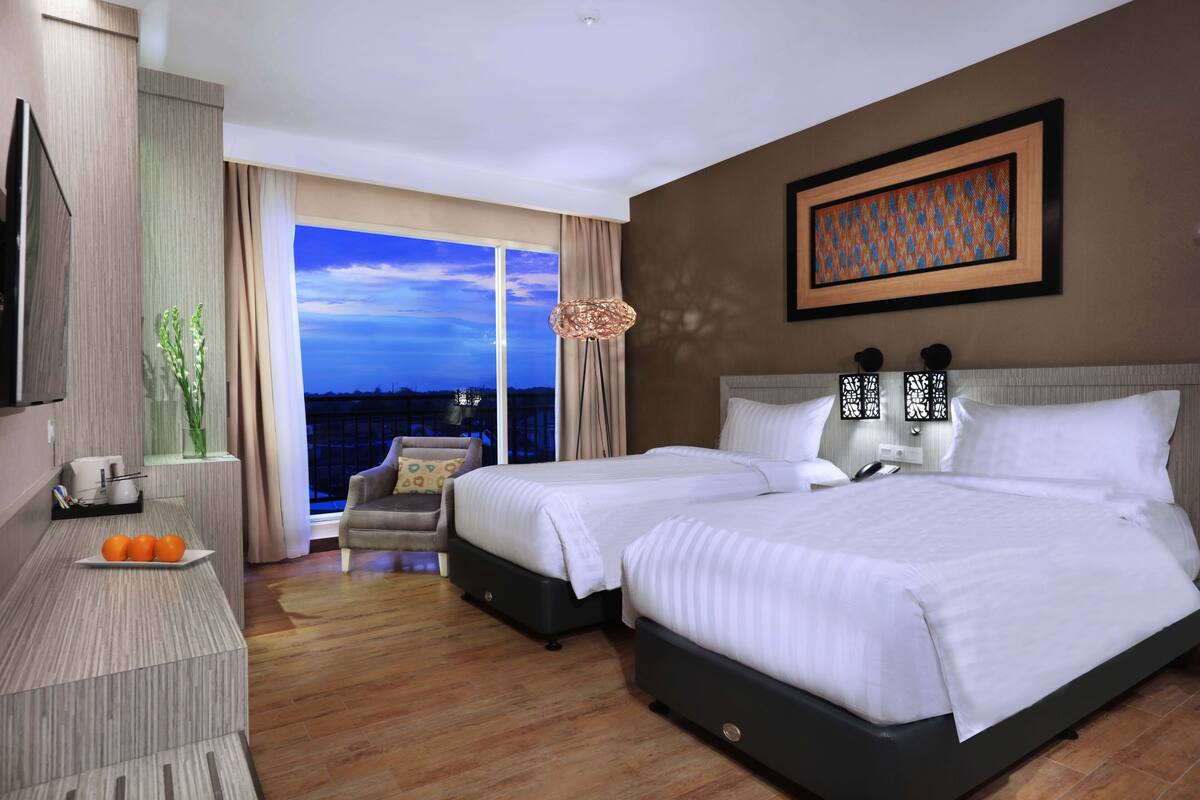 A hotel room with two beds, a TV, a desk with oranges, a chair, and a view of the city.