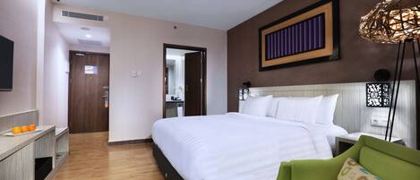 A hotel room with a bed, a green armchair, a bedside table, a TV, and a wooden floor.