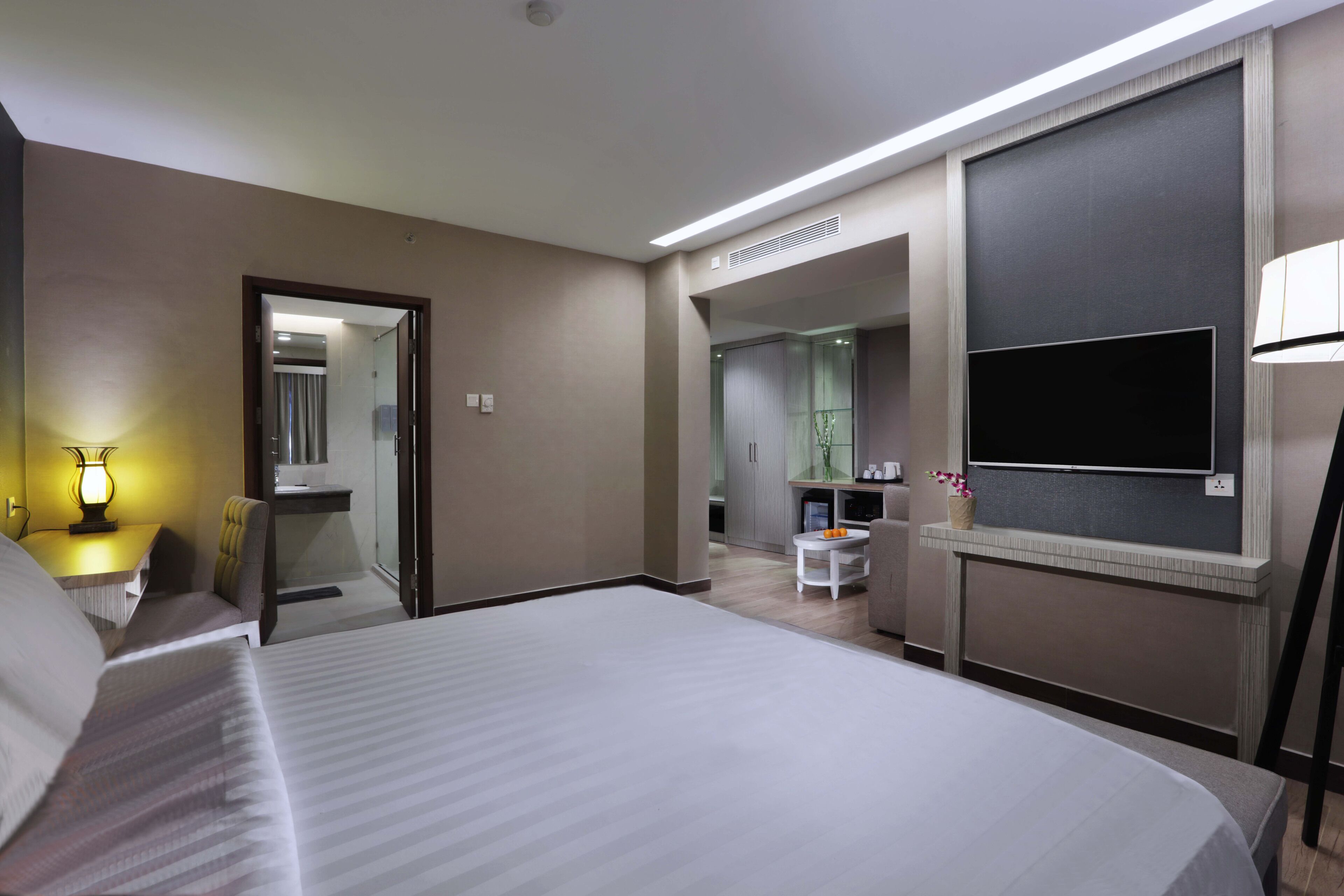A hotel room with a large bed, a flat-screen TV mounted on the wall, a desk with a lamp, and a small table with a vase of flowers.