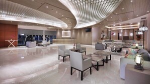 Un'ampia hall dall'arredamento moderno, con soffitto curvo e motivo geometrico, zona relax con divani e tavoli, e un grande schermo televisivo.
