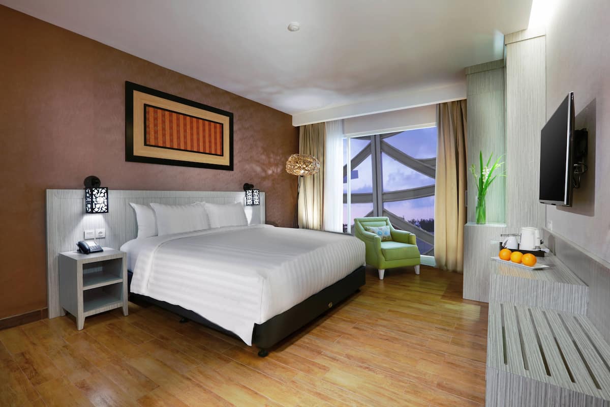 A modern hotel room with a large bed, a green armchair, a TV, and a window with a city view.