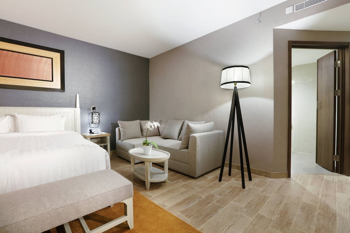 A hotel room with a bed, a sofa, a small round table with a plant, a floor lamp, and a door leading to another room.