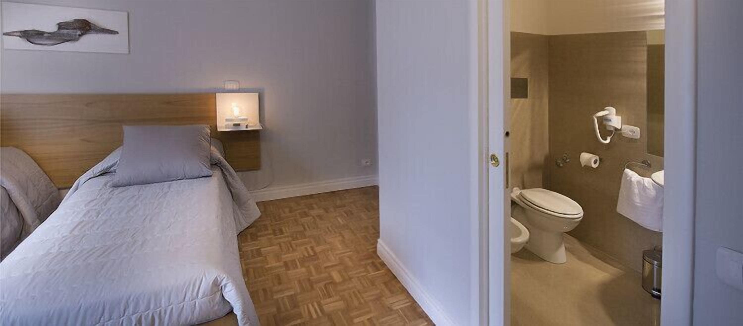 Ein Schlafzimmer mit Bett, Nachttisch mit Lampe sowie ein Badezimmer mit Toilette und Handtuchhalter.