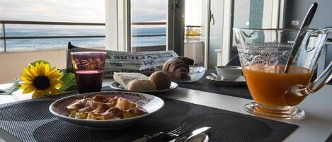 A breakfast table with a plate of food, a glass of orange juice, a newspaper, and a view of the ocean.