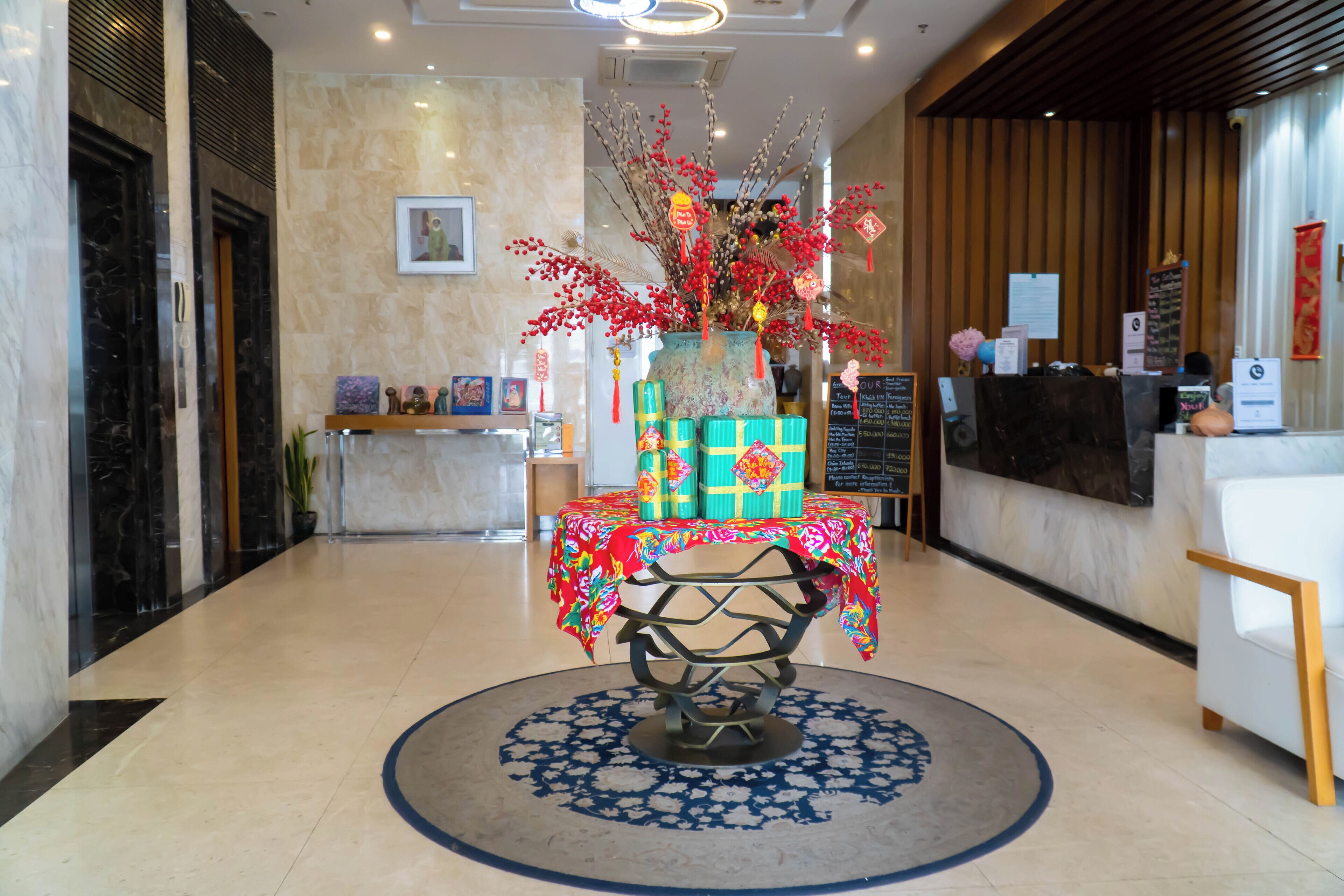 A hotel reception area with a reception desk, a decorative vase with red branches, and a round table with a patterned tablecloth.