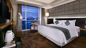 A modern hotel room with a large bed, a flat-screen TV, a desk with a lamp, and a view of the city through large windows. - Aston Pasteur (Bandung)