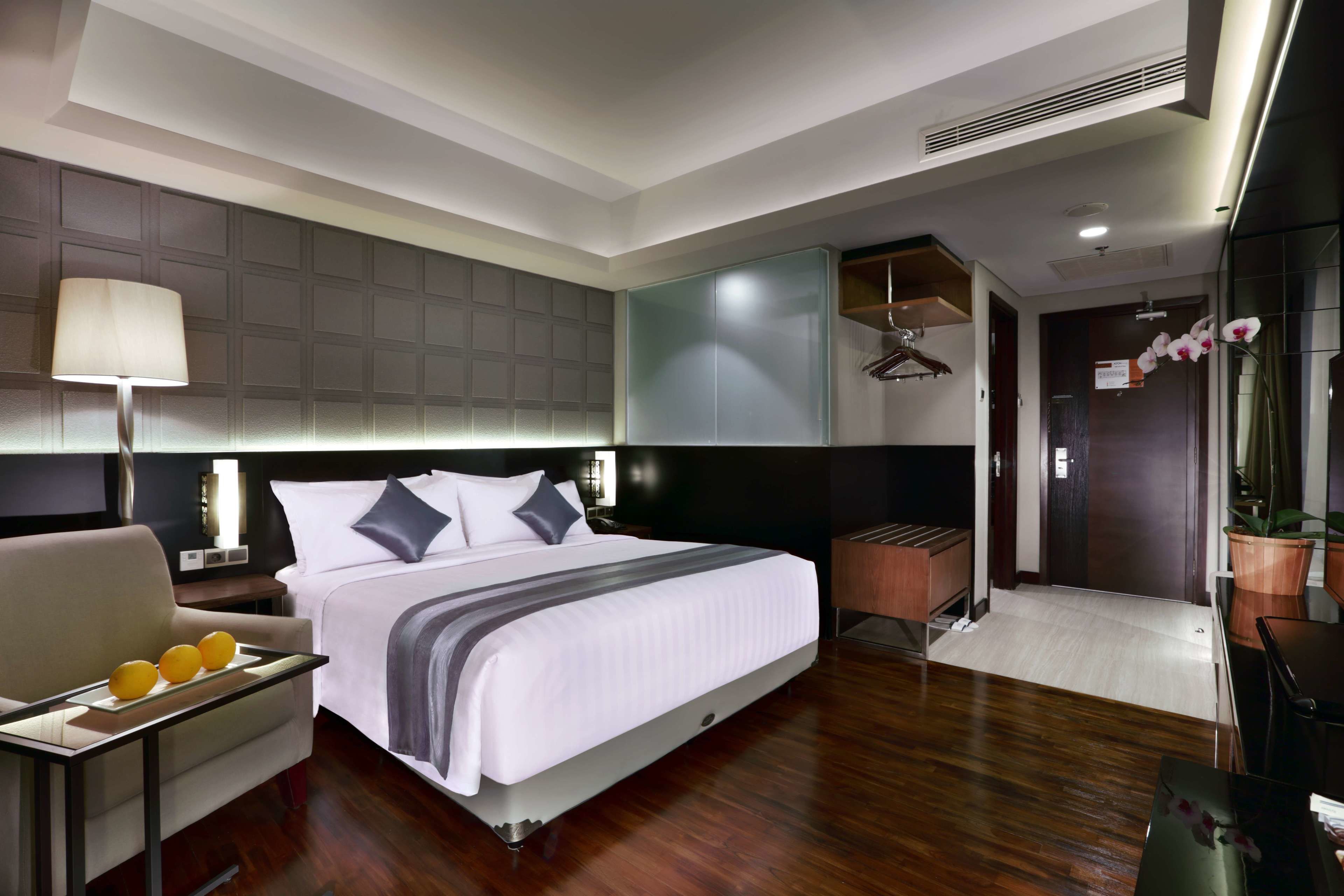 Una habitación de hotel moderna con una cama grande, una mesita de noche con lámpara, una silla y una pequeña mesa con naranjas.