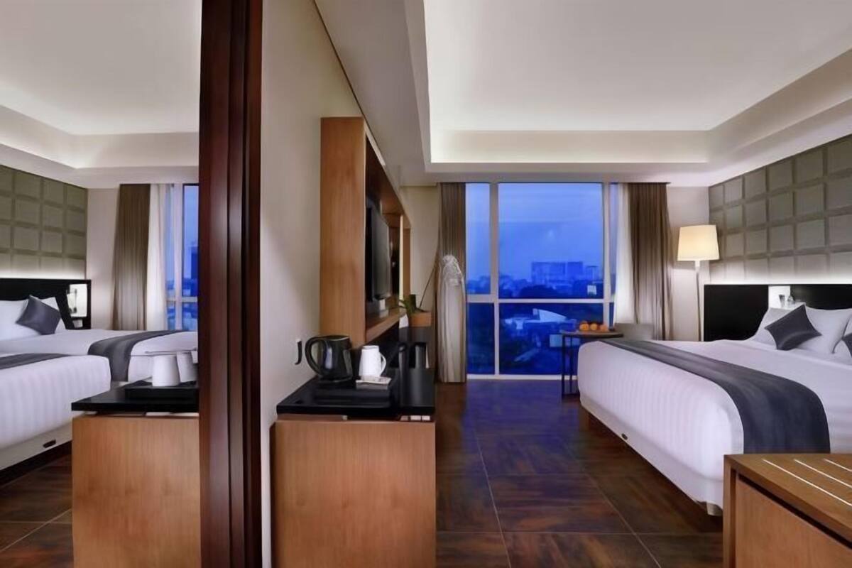 A hotel room with two beds, a coffee table, a desk, and a large window with a city view.