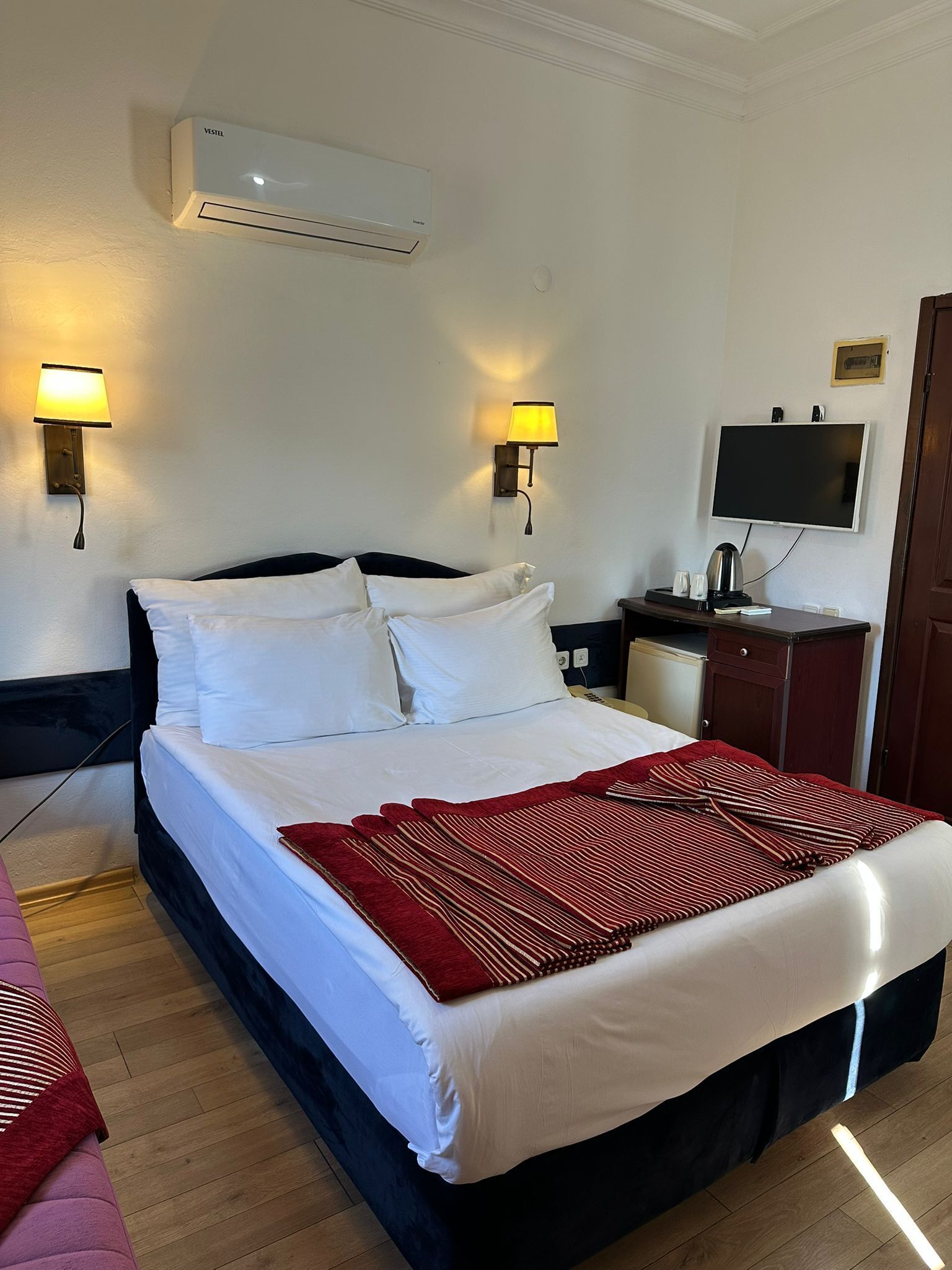 A hotel room with a bed, bedside lamps, a flat-screen TV, an air conditioner, and a wooden floor.