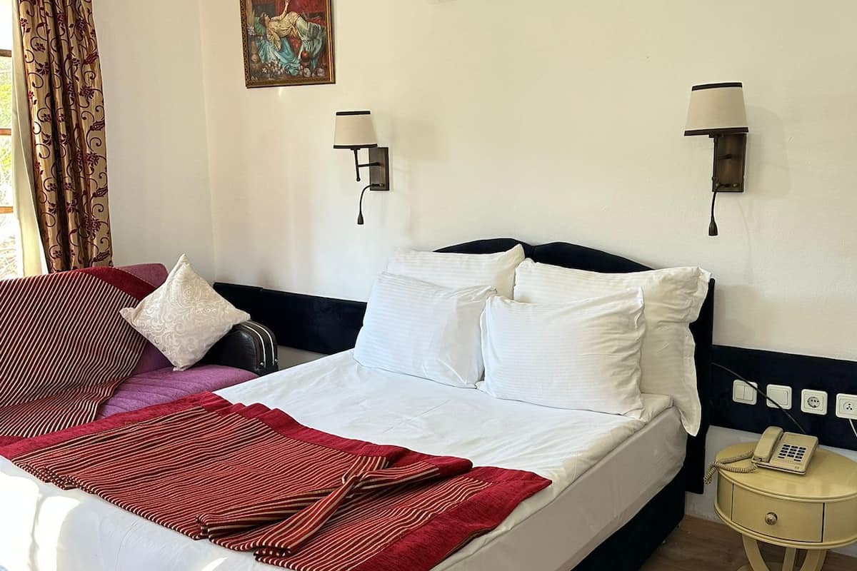 Habitación de hotel con cama, lámparas de noche, aire acondicionado, un cuadro y un teléfono sobre una mesita.
