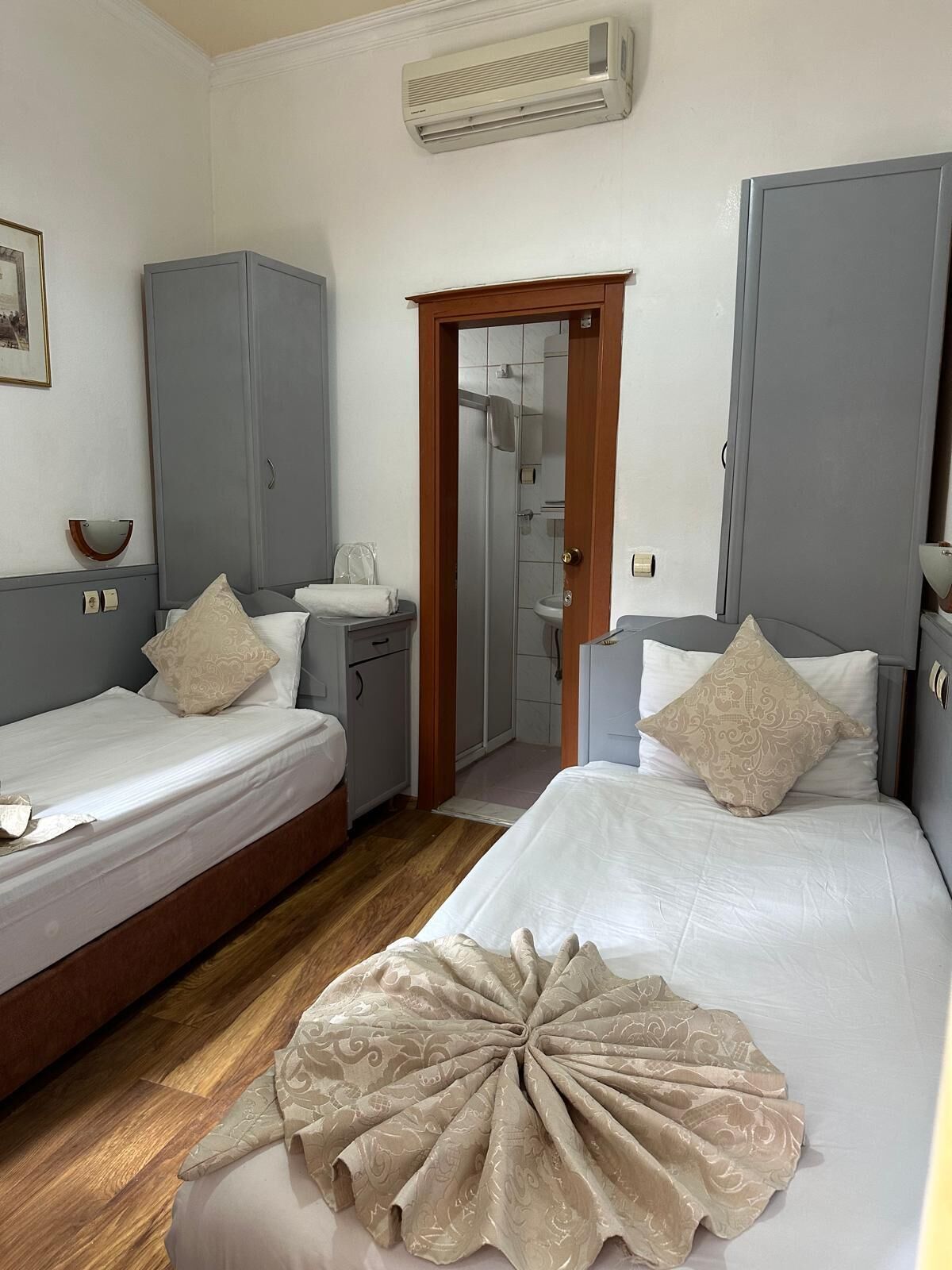 A hotel room with two beds, a wardrobe, a bathroom visible through a door, and an air conditioning unit on the wall.