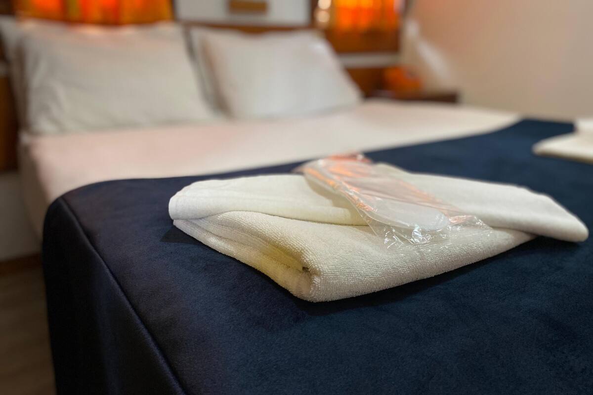 Una cama bien hecha con una colcha azul y almohadas blancas, una toalla blanca doblada y una bolsa de plástico sobre la cama.