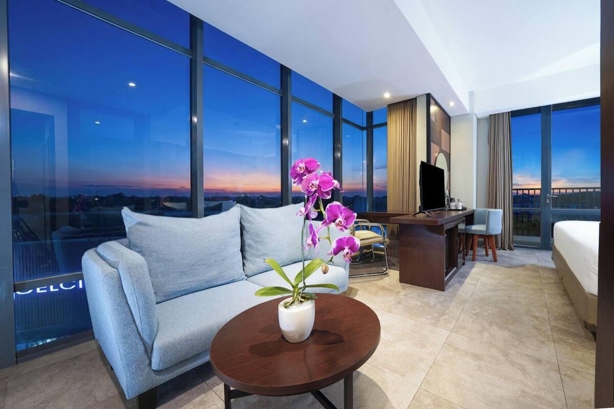 A modern hotel room with a large window, a sofa, a desk, a chair, and a bed.