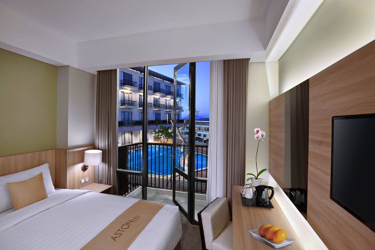 A hotel room with a bed, a desk with a plate of fruit, a TV, and a view of a pool and building.