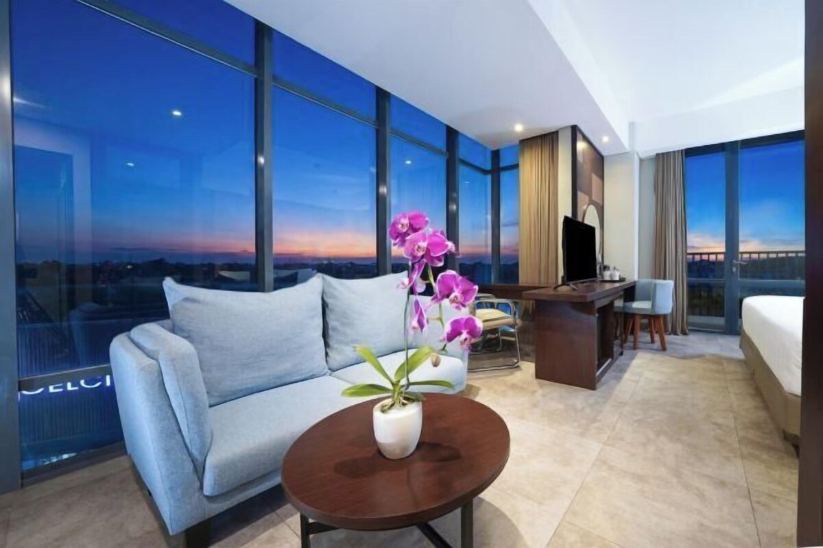 A modern hotel room with a large window, a sofa, a round table with a plant, a desk, and a bed.