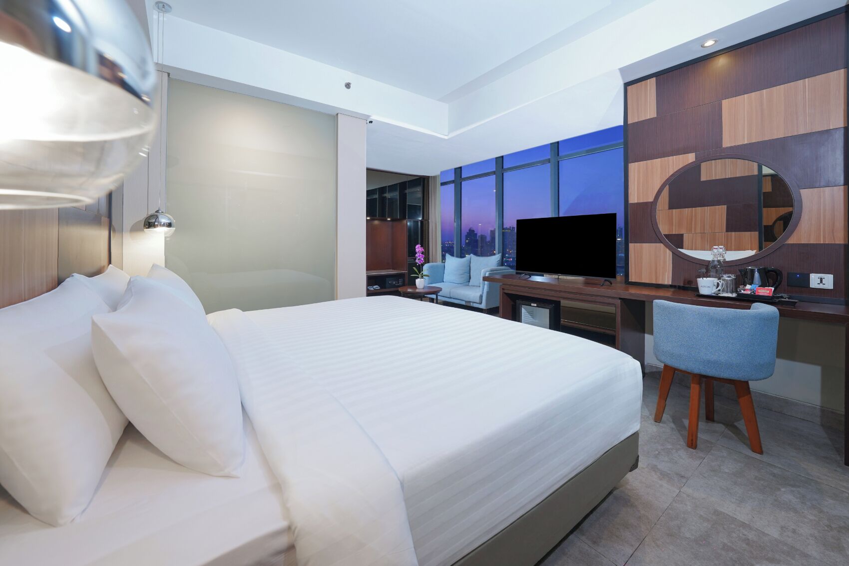 A modern hotel room with a large bed, a desk with a TV, and a view of the city at dusk.