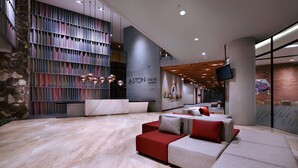 A modern hotel lobby with a large reception desk, a seating area with red and grey sofas, and a stone wall on one side. - Aston Inn Mataram (Mataram)