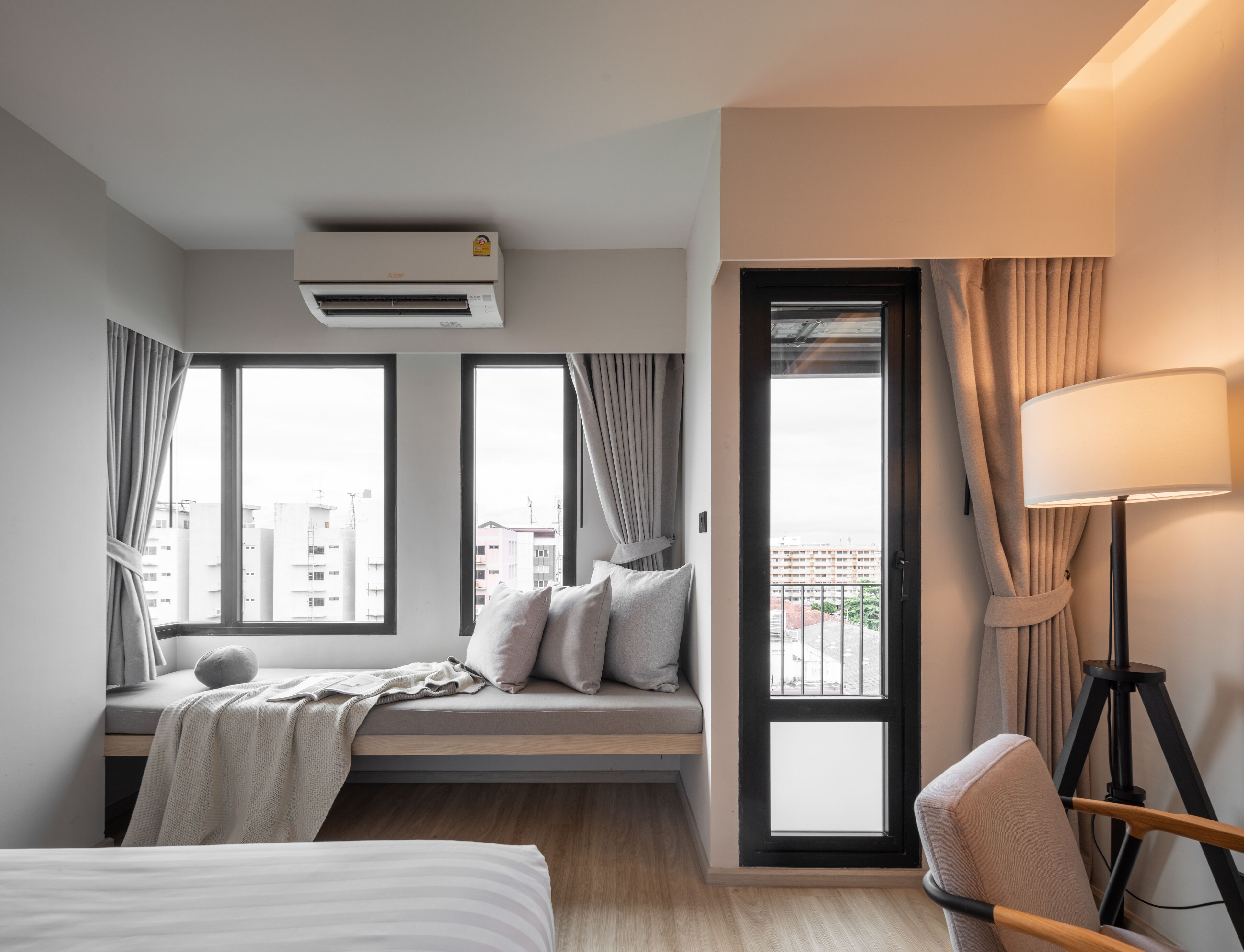 Habitació d'hotel moderna amb un llit, zona de seients amb coixins, escriptori, làmpada i grans finestres amb cortines.