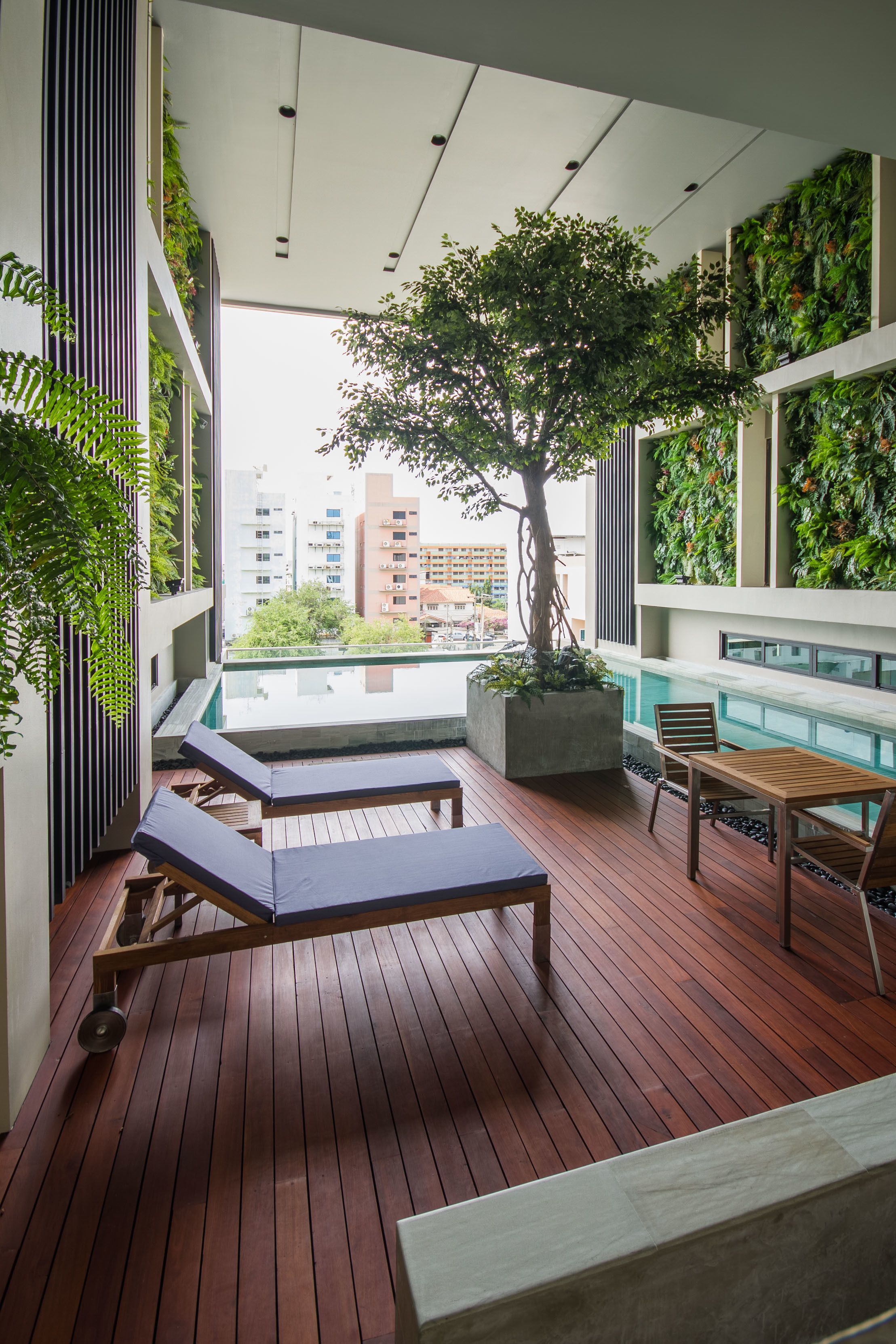 A modern outdoor lounge area with wooden flooring, lounge chairs, a table, and a large tree.