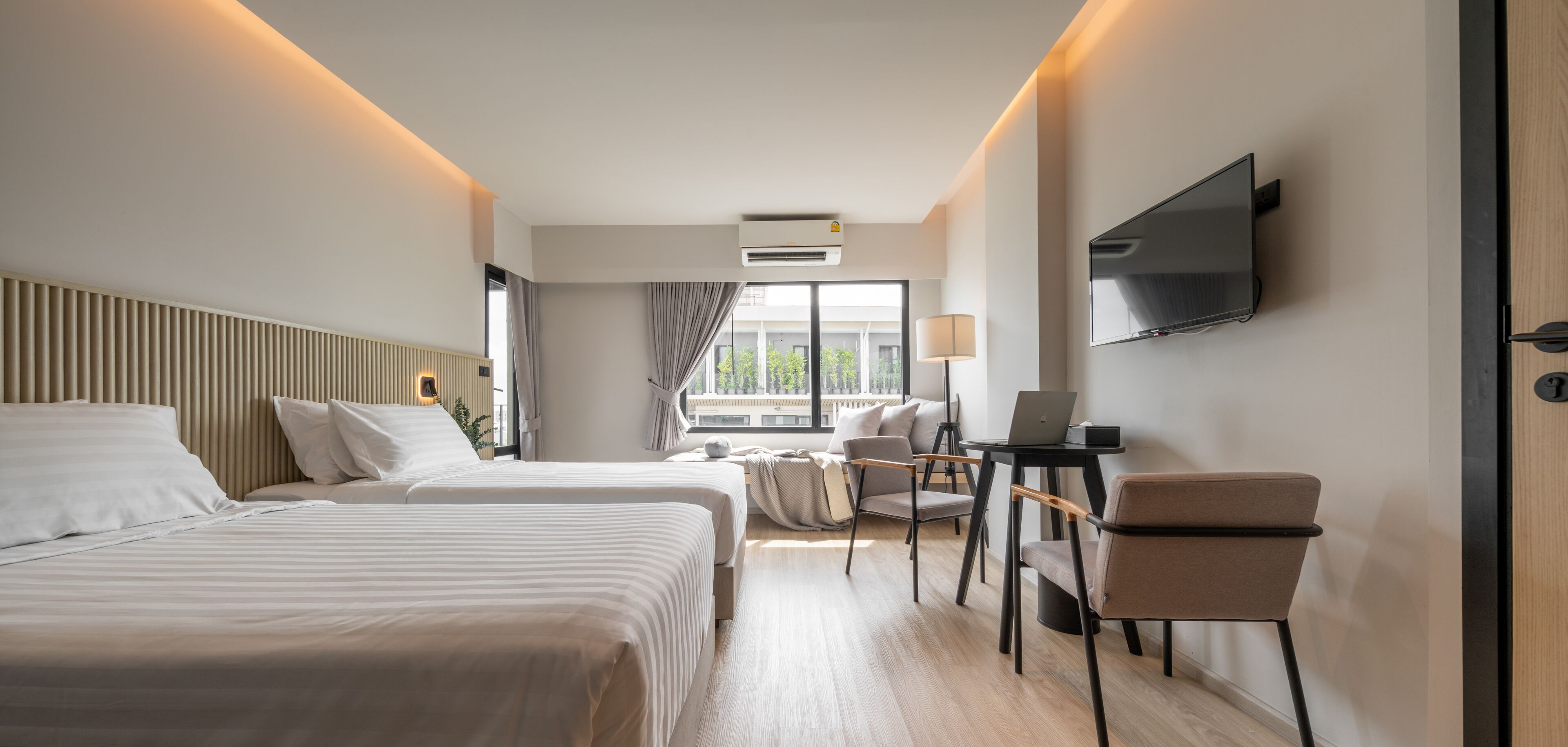 A modern hotel room with two beds, a desk with a laptop, a TV, and a large window with a view of buildings.