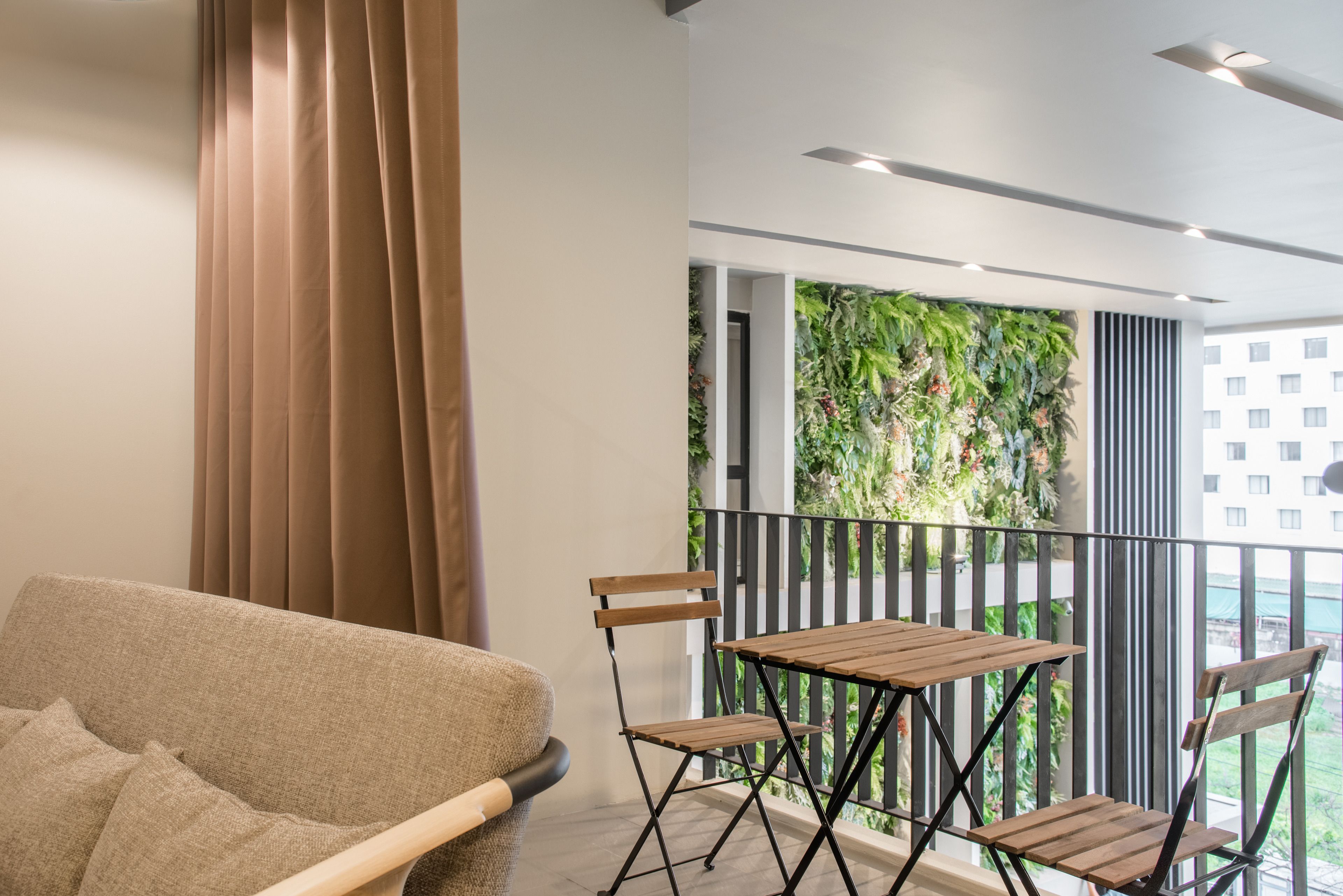 A balcony with a view of a building and a green wall. There is a sofa and two wooden chairs.