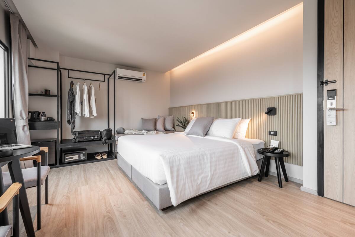 A modern hotel room with a large bed, a desk with a computer, a chair, and a small table with a lamp.