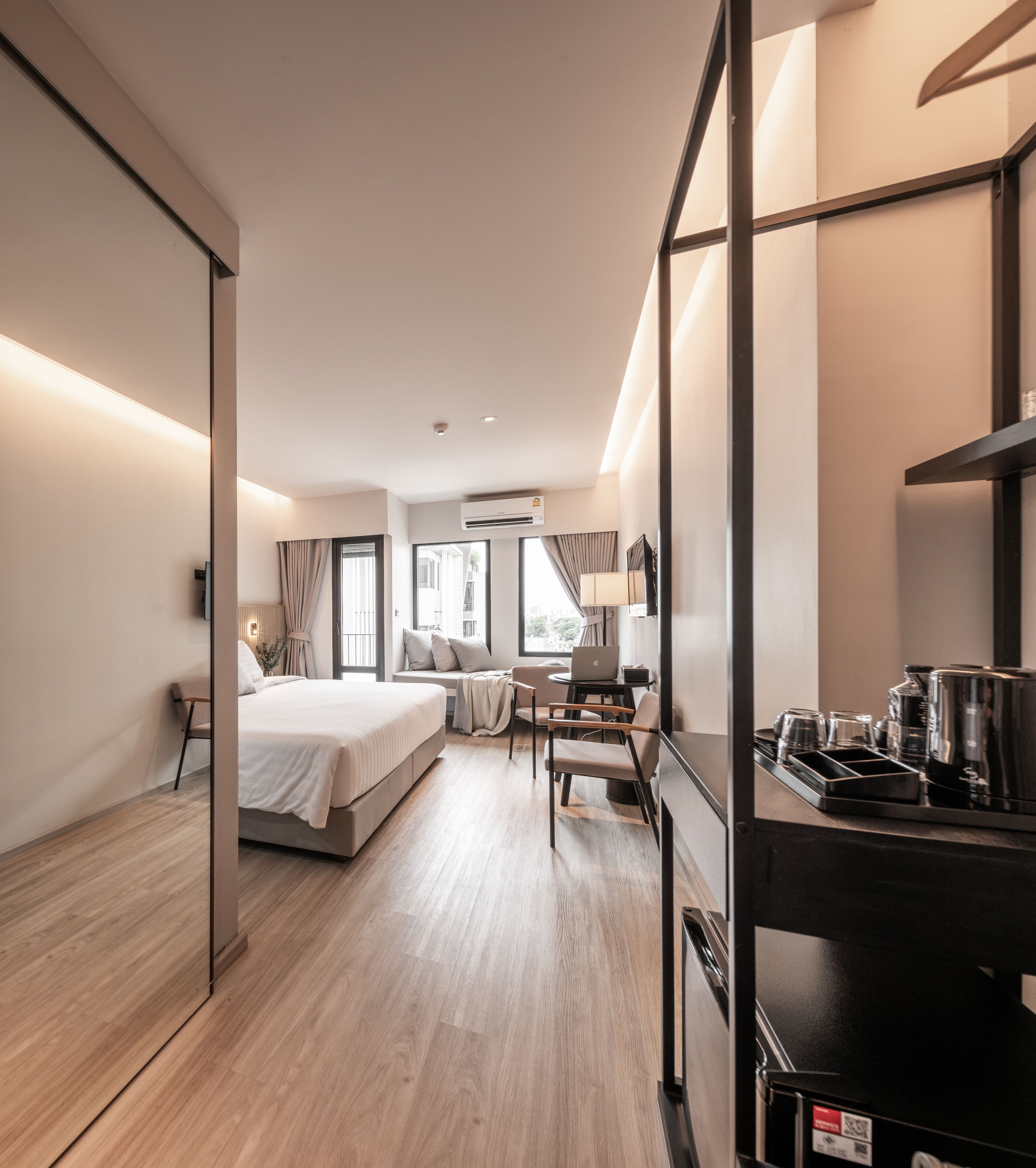 Una habitación de hotel moderna con cama, escritorio con computadora portátil, cafetera y vista de la ciudad a través de amplias ventanas.