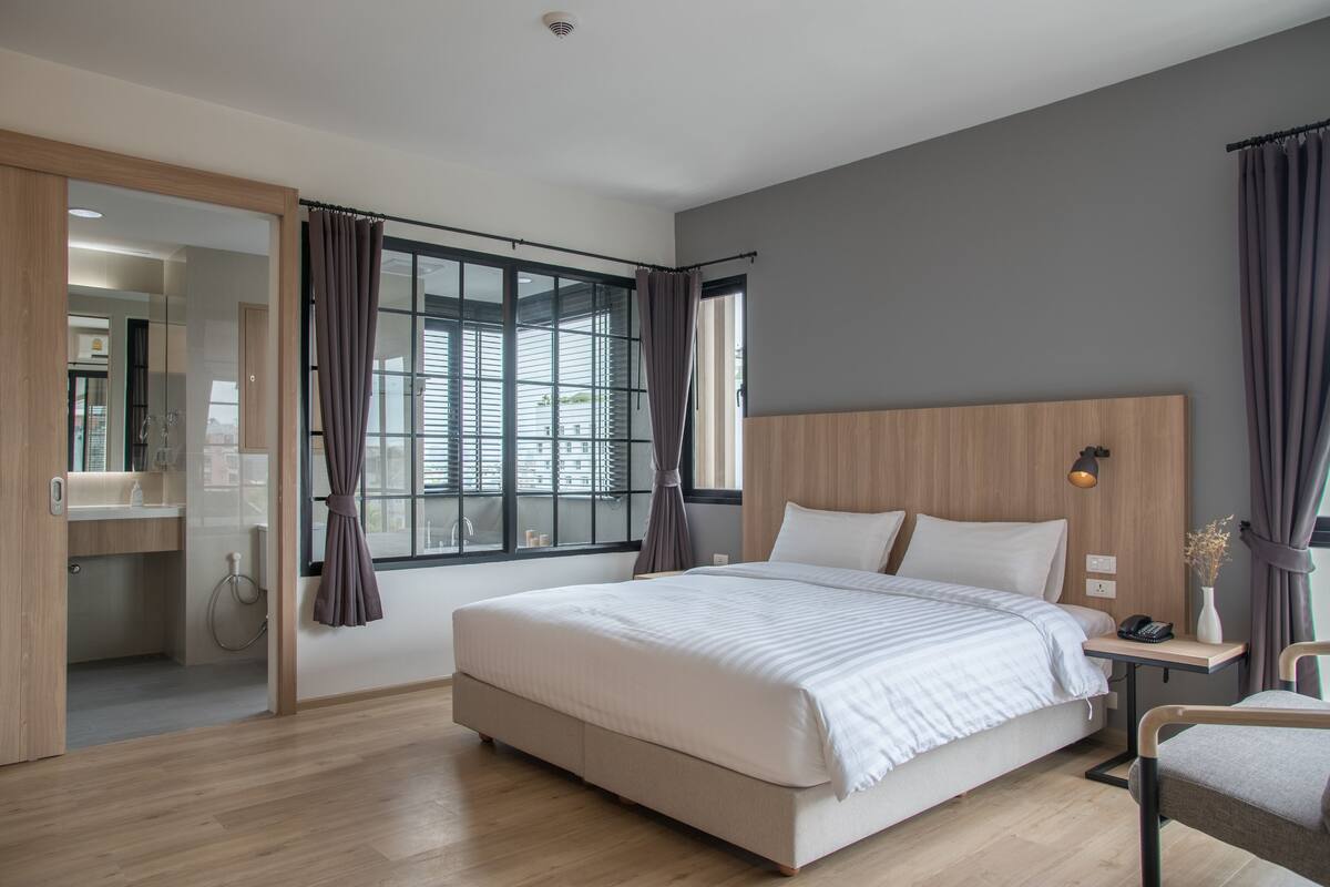 A modern bedroom with a large bed, a wooden headboard, a bedside table with a lamp, and a view of the bathroom.