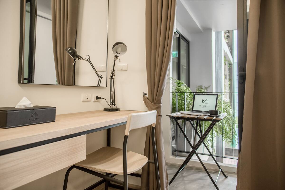 A modern hotel room with a desk, chair, and a small table with a laptop and a bowl. A mirror and a lamp are on the wall.