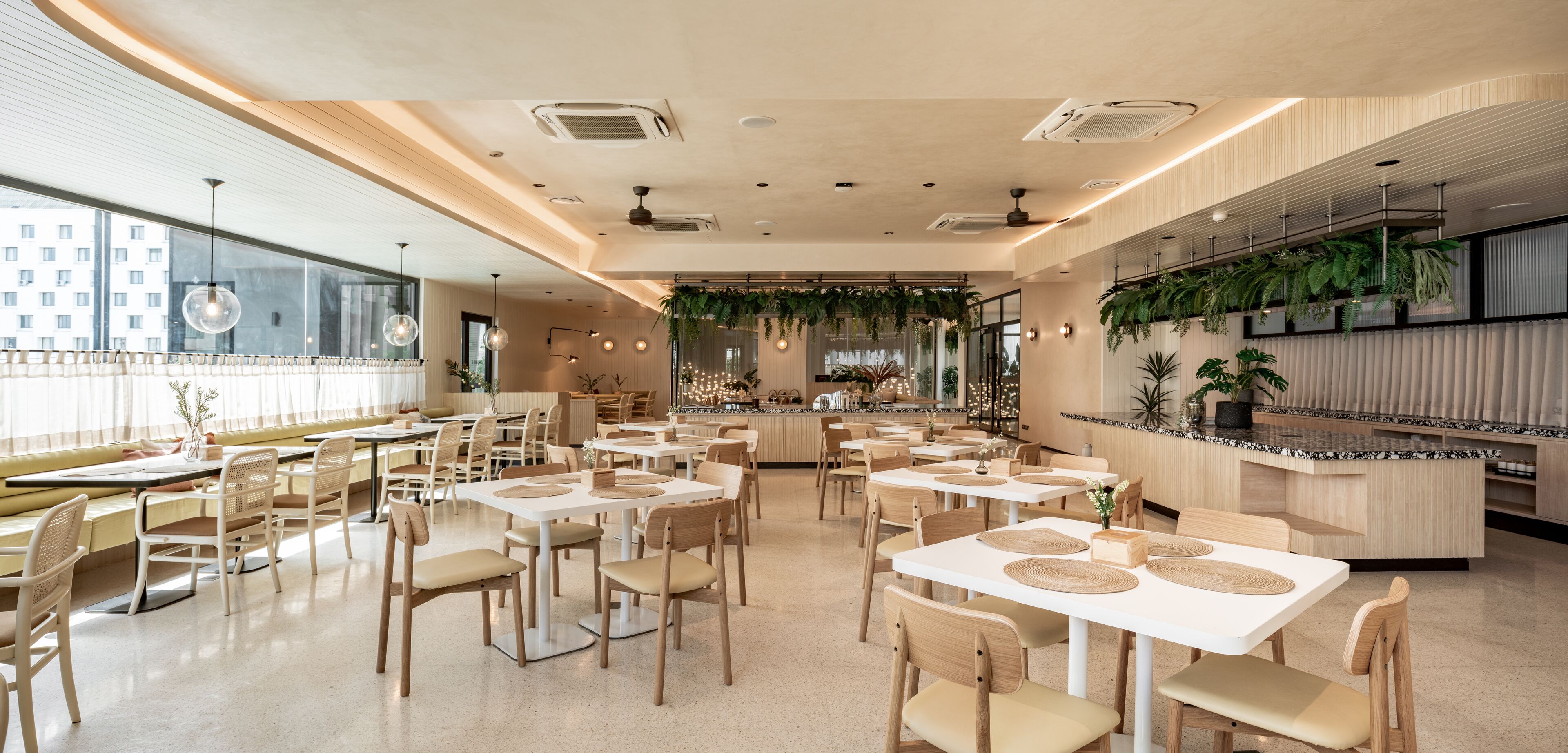 A modern restaurant with white tables and chairs, a bar area with plants, and large windows.